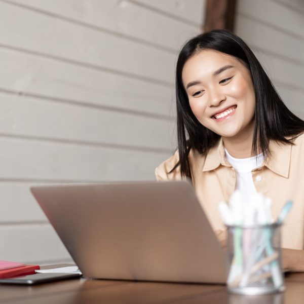 Smiling asian girl looks at laptop screen, works from home, listens webinar or attends online lecture, university classes on remote.